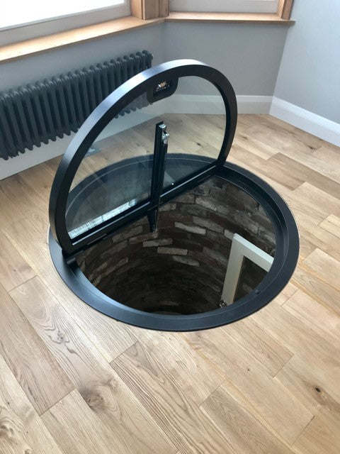 Round glass access hatch on a wooden floor with a view into deep well.
