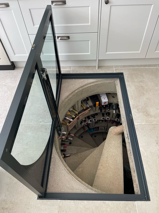 A hinged walk-on glass cellar door in the open position, with gas struts visible, revealing a spiral concrete staircase and wine cellar below.